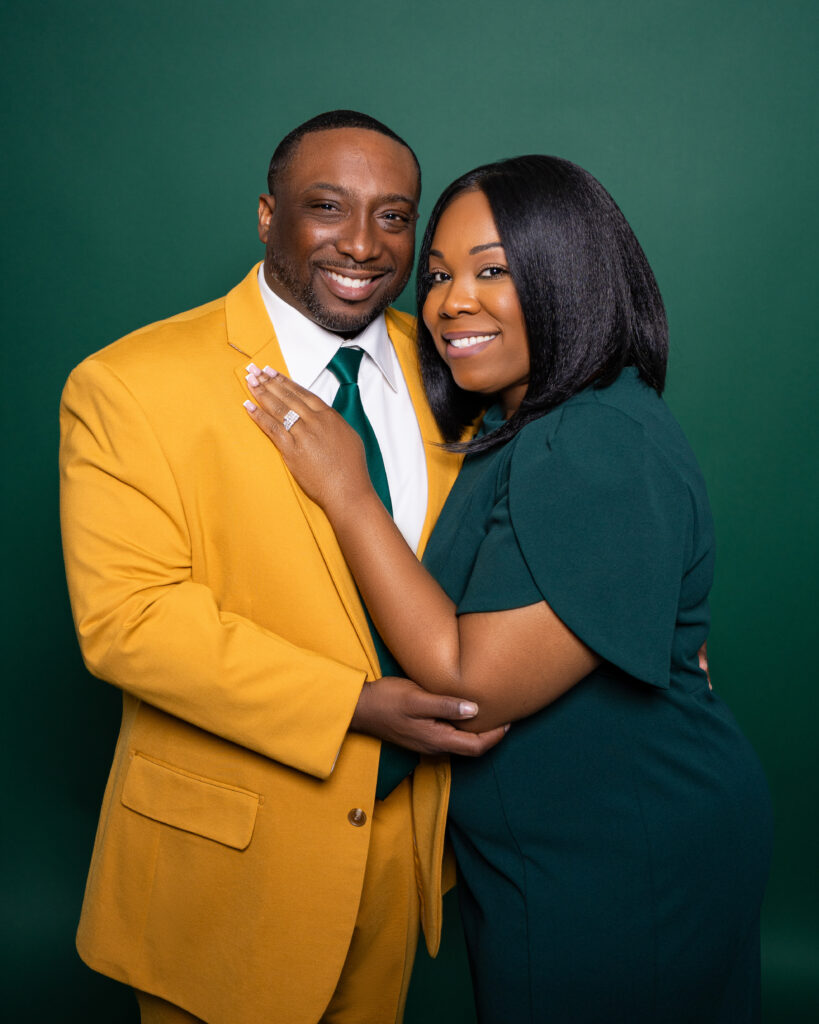 A couple embraces happily, dressed in yellow and green against a green backdrop.