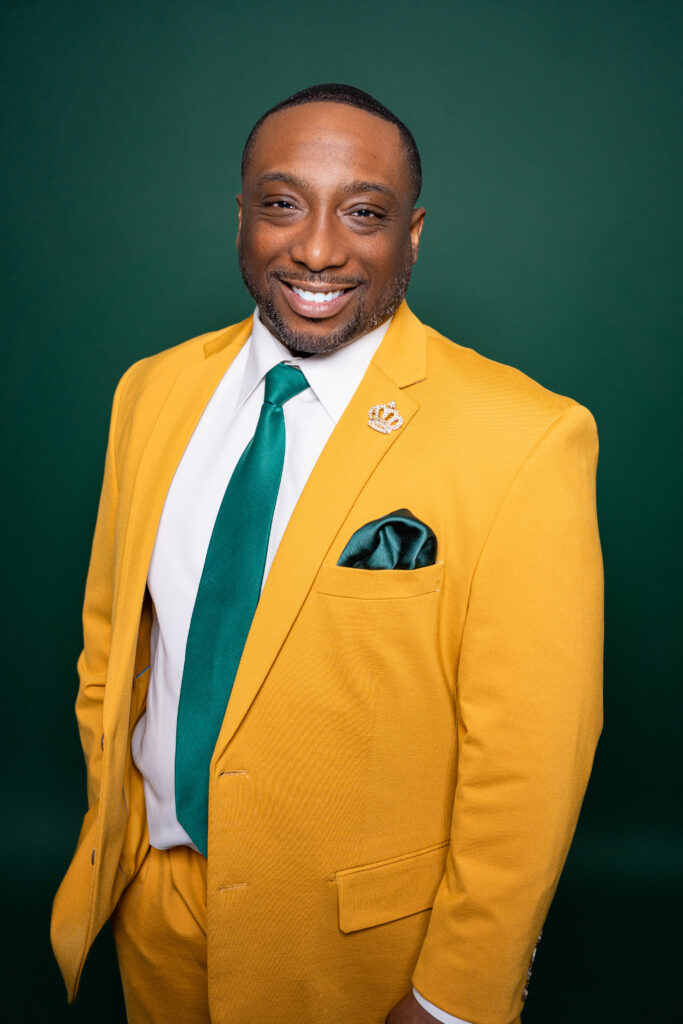 A man smiles confidently wearing a yellow blazer and green tie against a green background.